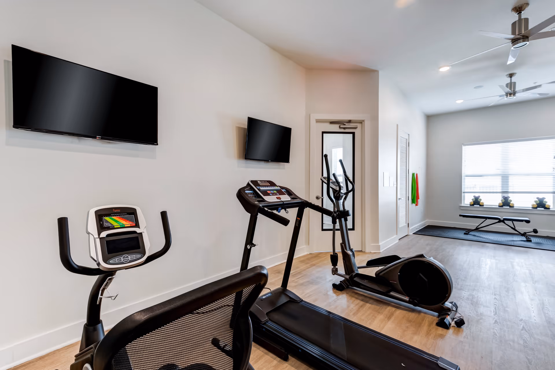 A fitness room with exercise equipment including a treadmill, an elliptical machine, and a stationary bike. Two flat-screen TVs are mounted on the wall above the equipment. In the background, there is a bench, some small weights on the windowsill, and resistance bands hanging on the wall. The room has light-colored walls, wood flooring, ceiling fans, and a window letting in natural light.