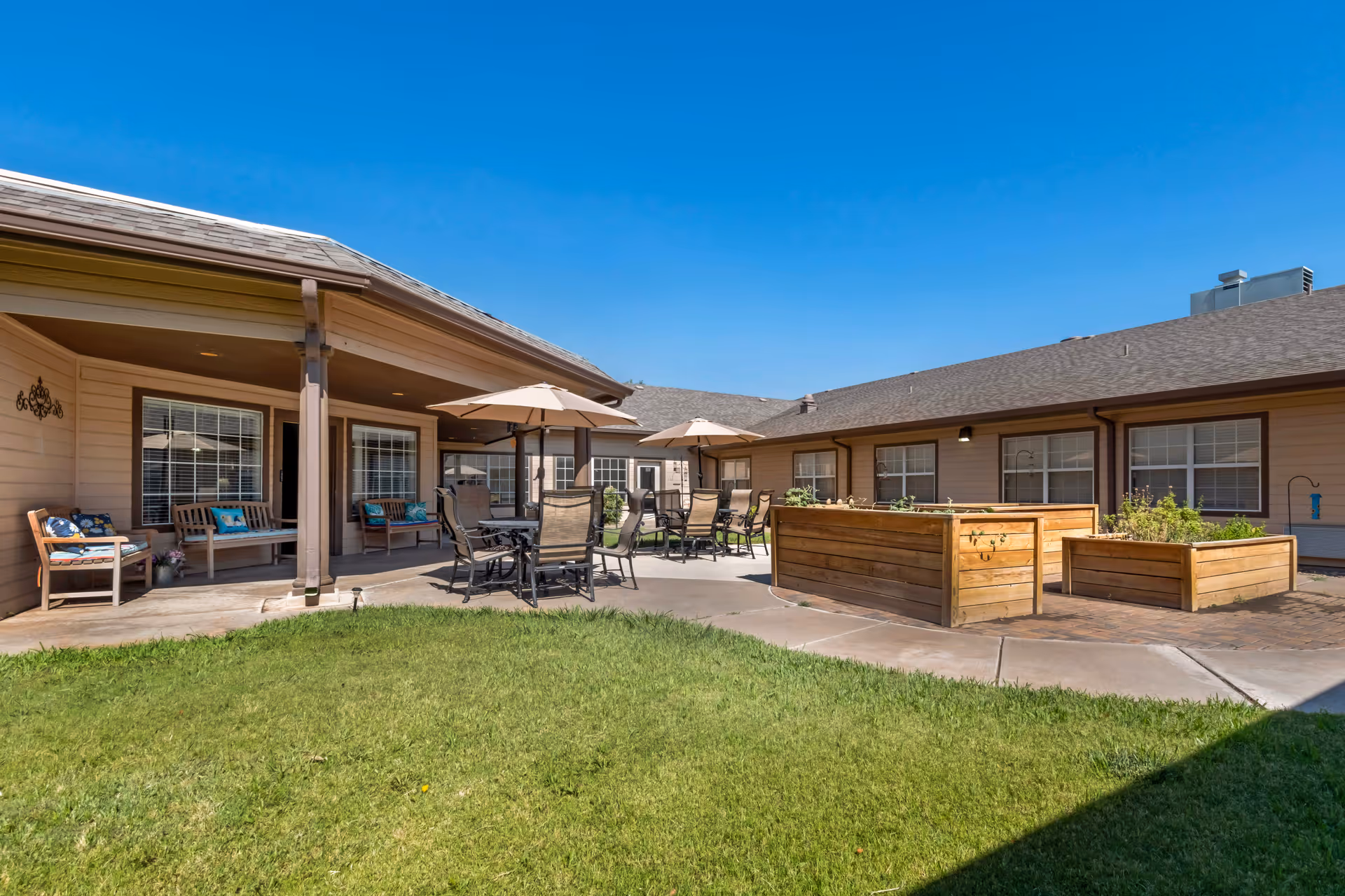 Outdoor courtyard with patio tables and umbrellas, benches, and raised wooden planters beside a single-story senior living building.