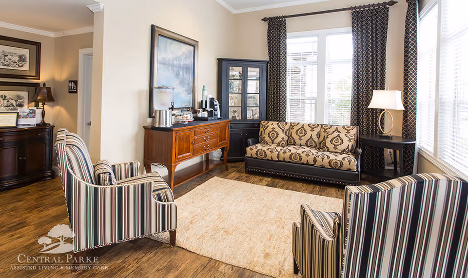 A cozy living room area with a patterned sofa, two striped armchairs, a wooden sideboard with a coffee station, a black display cabinet, a round side table with a lamp, large windows with patterned curtains, and a beige rug on wooden flooring.