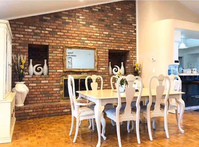 A white dining table with six chairs set on a parquet floor in front of a brick fireplace wall decorated with vases and a mirror.