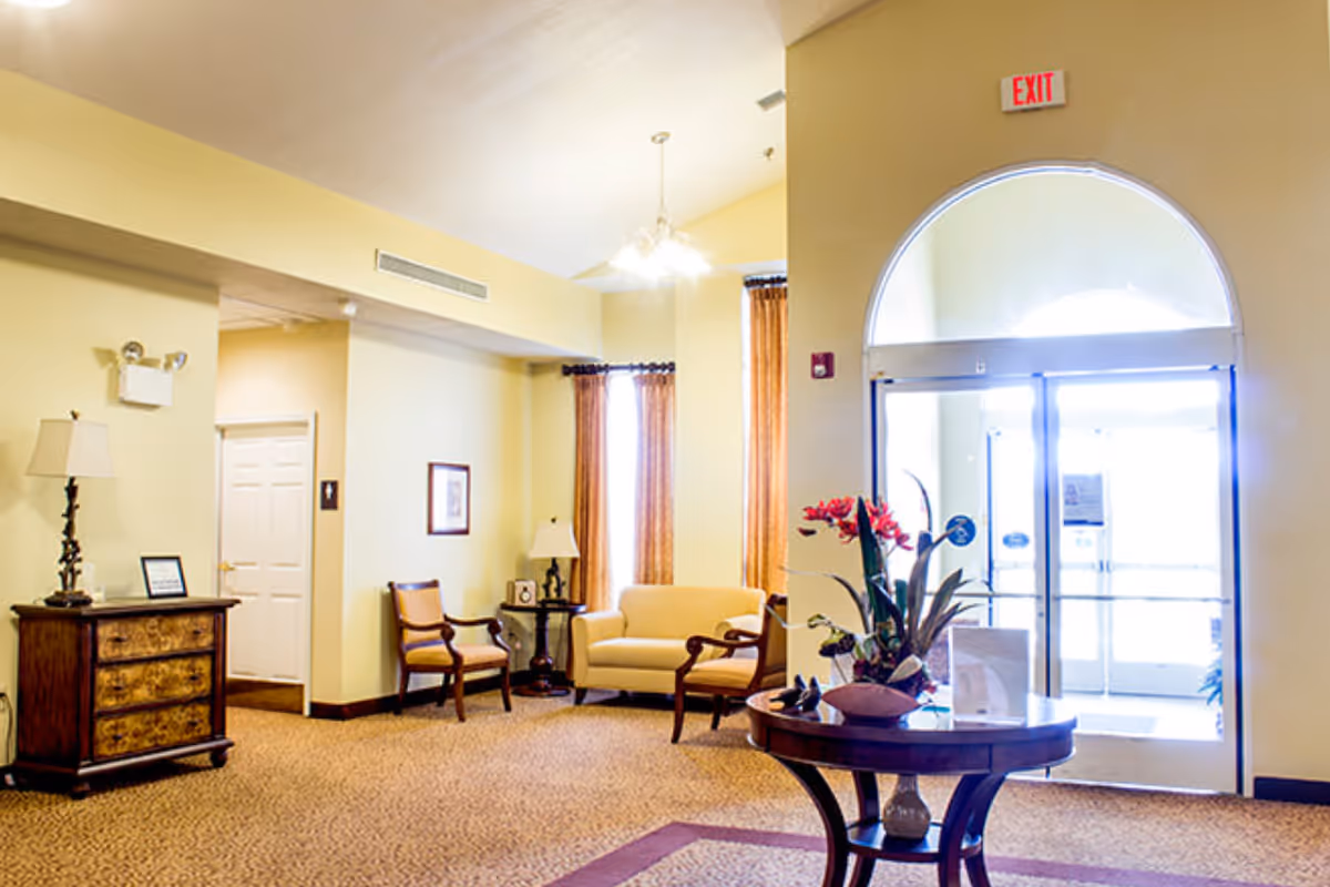 A bright and welcoming lobby area with beige walls and carpet. There is a round wooden table with a flower arrangement in the center of the room. To the left, there is a wooden chest of drawers with a lamp and framed picture on top. Two armchairs and a small sofa are arranged near two tall windows with brown curtains. A glass door with an arched window above it is visible on the right side, with an exit sign above the door.