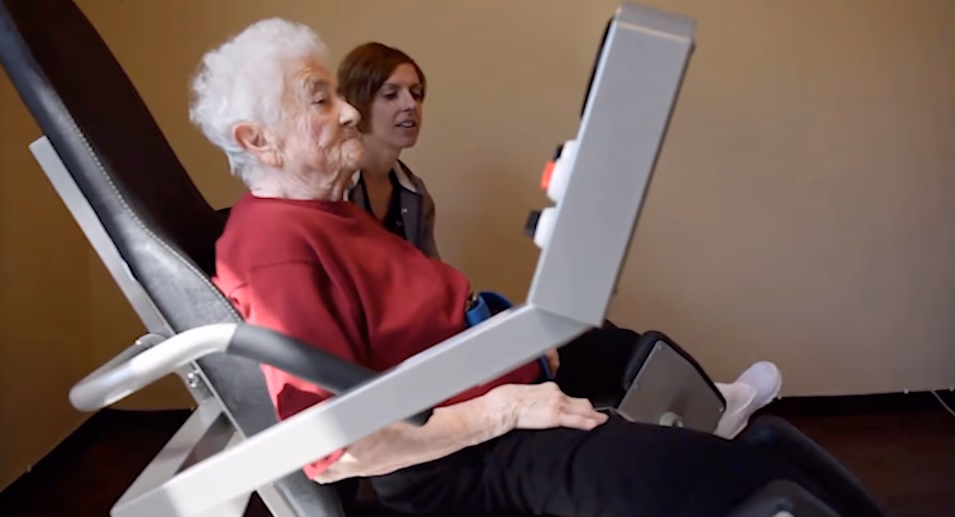 An elderly woman in a red sweater is seated on a piece of exercise equipment, with a younger woman beside her providing assistance or support in a room with plain beige walls.