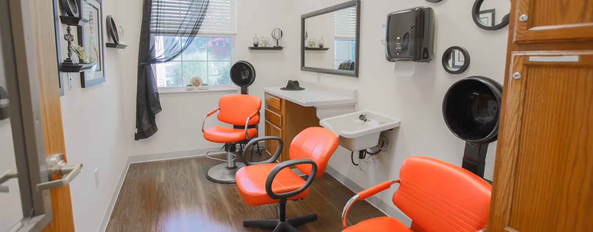 A small hair salon room with three bright orange salon chairs, two black hair dryers, a white sink, a large mirror above a wooden cabinet, and a window with blinds and a sheer black curtain.