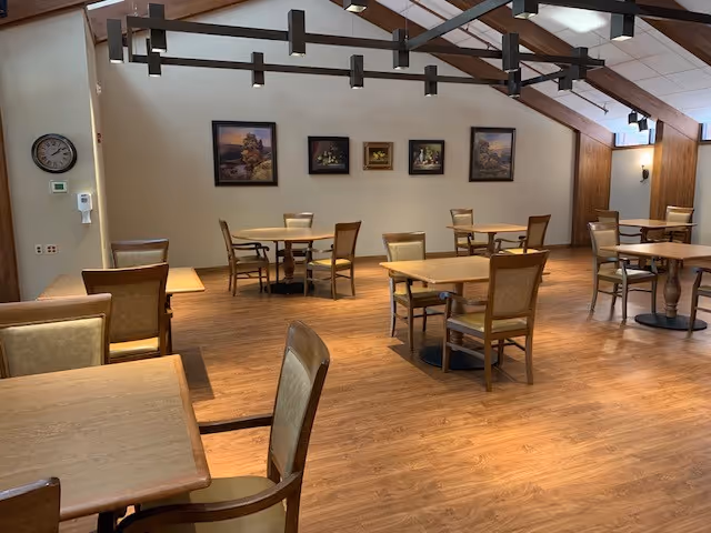 A spacious dining room with wooden floors and several square wooden tables, each surrounded by four chairs with cushioned seats and backs. The walls are decorated with framed paintings, and a large modern chandelier hangs from the ceiling. A clock and a hand sanitizer dispenser are mounted on one wall.