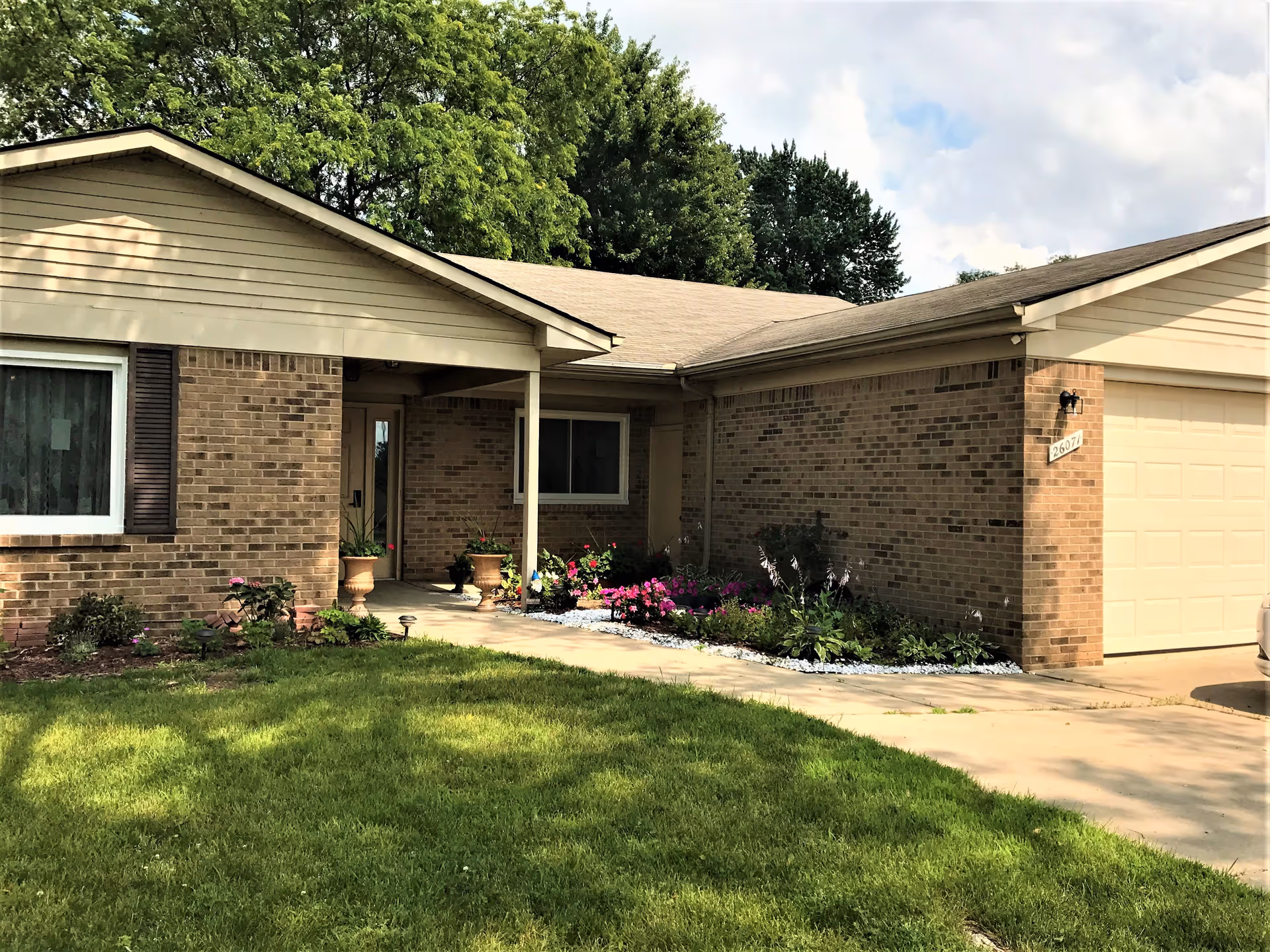 Front exterior view of a single-story brick house with a beige roof, a garage on the right, a walkway leading to the front door, and a garden with flowers and plants along the walkway. Trees are visible in the background under a partly cloudy sky.