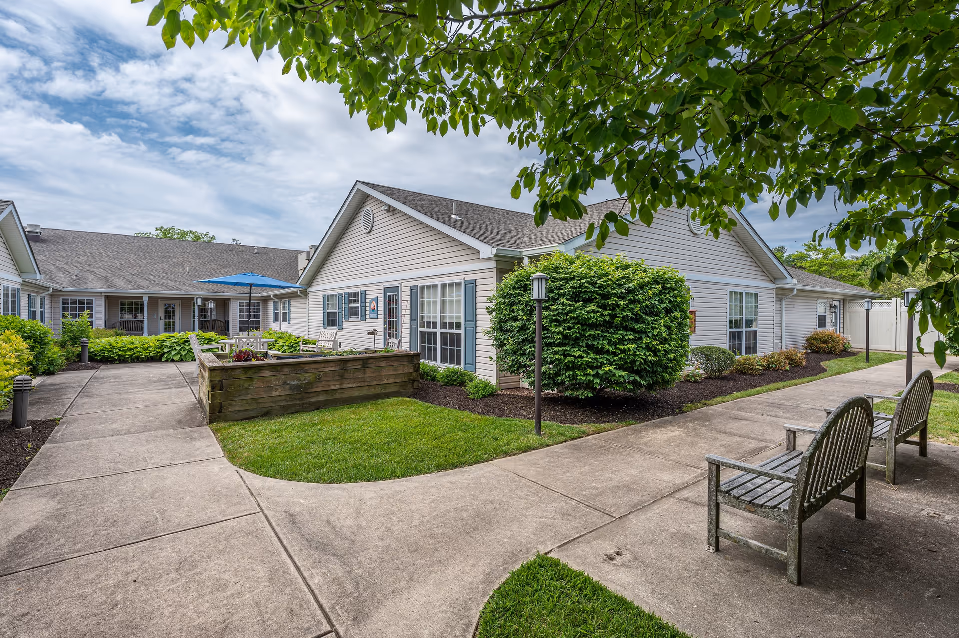 Outdoor courtyard area of Arden Courts - ProMedica Memory Care Community in Wilmington featuring a paved walkway, green lawn, bushes, benches, and a building with beige siding and multiple windows under a partly cloudy sky.
