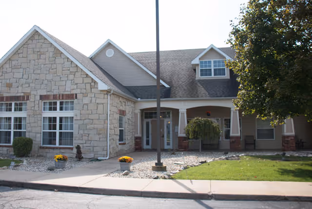 Exterior view of a single-story building with stone and siding walls, multiple windows, a covered entrance with columns, a small landscaped area with grass, rocks, and potted flowers, and a tree on the right side.