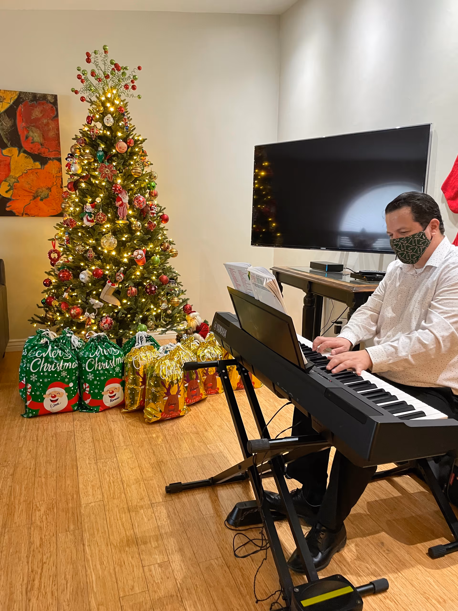 A masked man plays an electronic keyboard beside a decorated Christmas tree with wrapped gifts in a cozy living room.