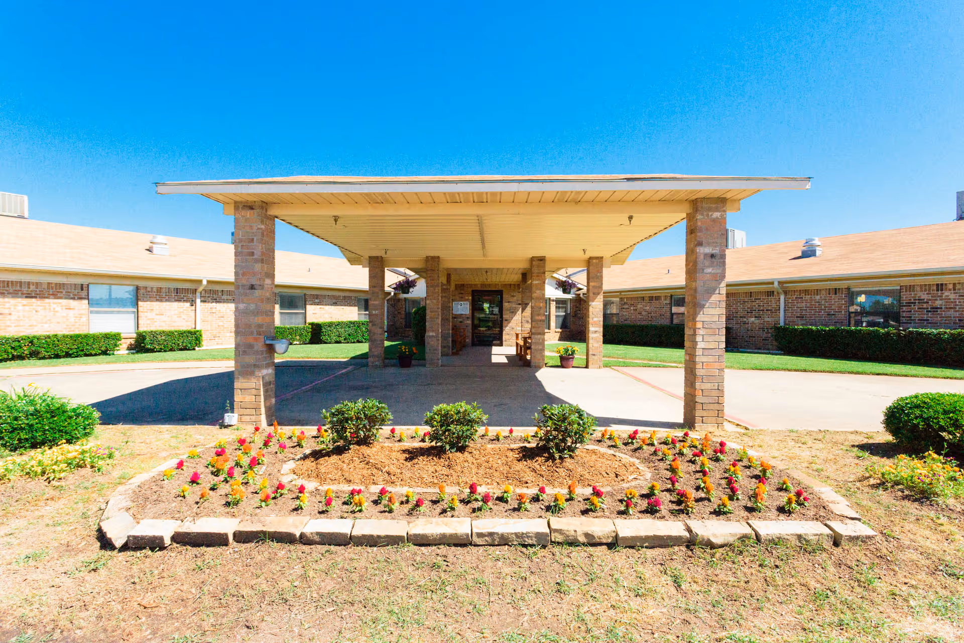 Entrance of a single-story brick building with a covered driveway supported by brick pillars, a flower bed with small plants and flowers in front, and a clear blue sky above.