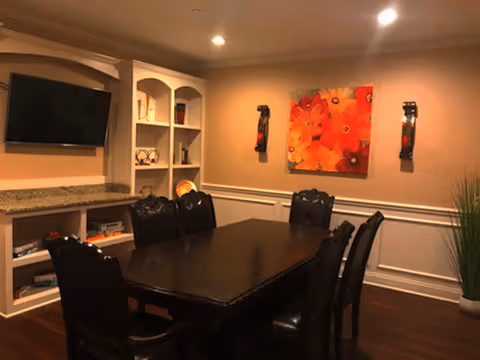 Dining room with a dark wood table and chairs, built-in white shelving and a wall-mounted TV, and a colorful orange floral painting on the wall.