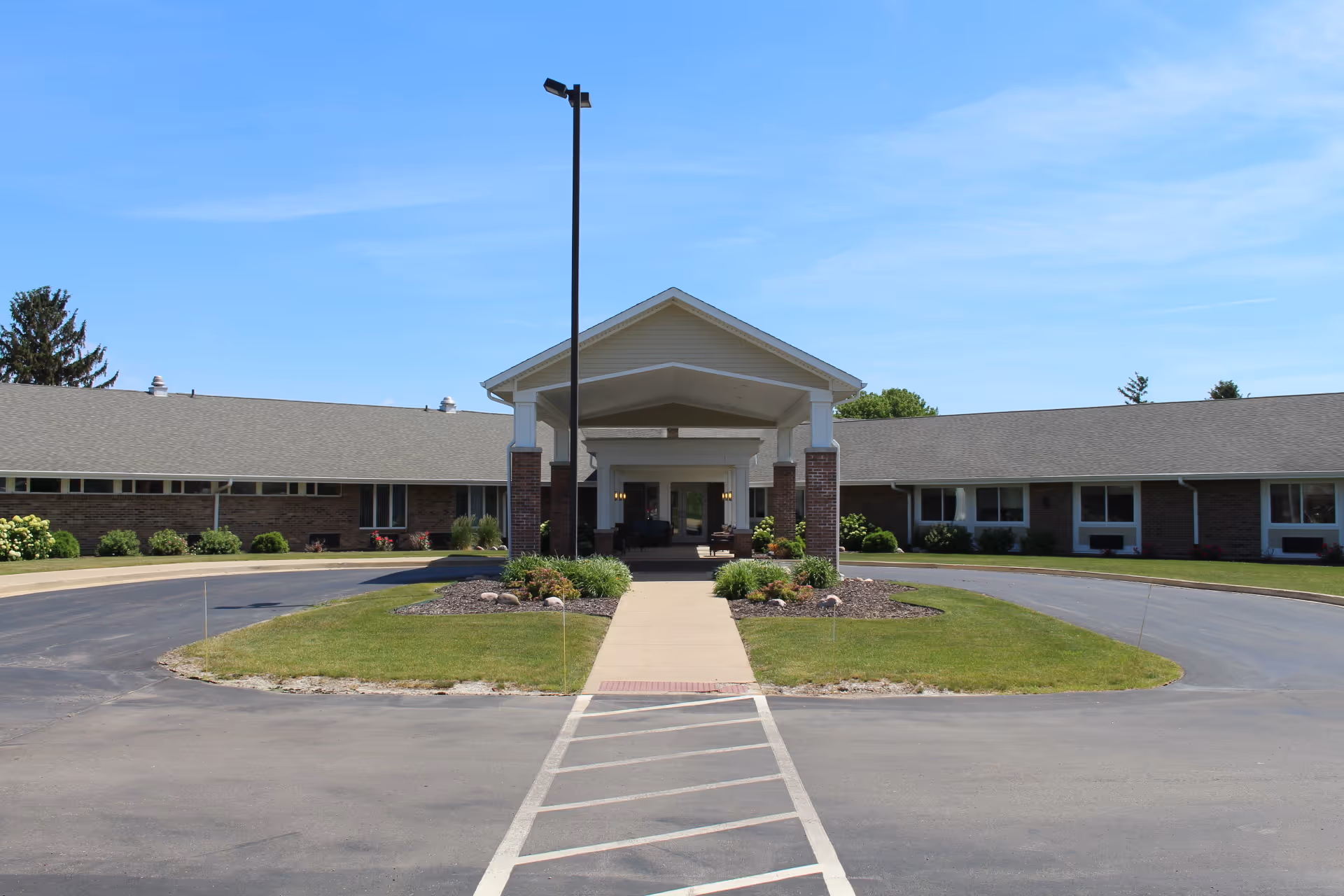 Front exterior view of a single-story brick building with a covered entrance supported by white columns. There is a paved driveway and a walkway leading to the entrance, surrounded by green grass and landscaped plants under a clear blue sky.