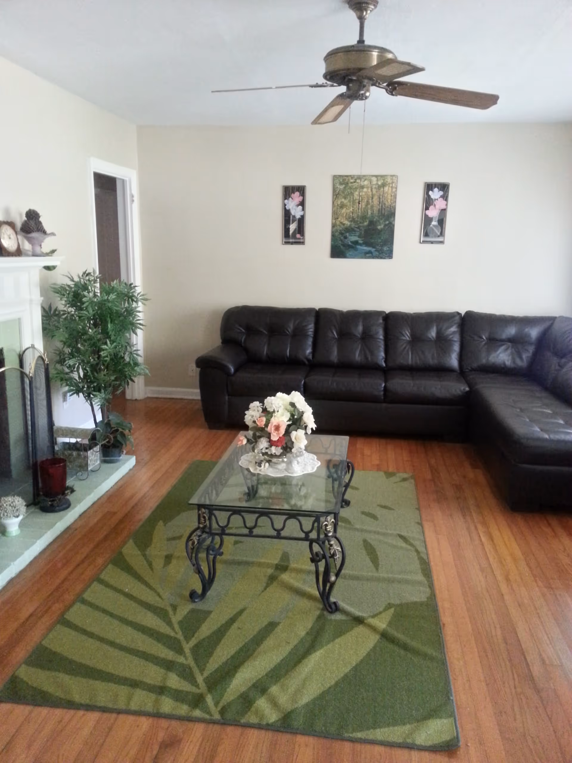 A living room with a large dark brown leather sectional sofa against a beige wall. Above the sofa are three framed pictures, one large central image of a forest and two smaller floral prints on either side. A glass coffee table with ornate black metal legs sits on a green rug with a leaf pattern. On the coffee table is a floral arrangement in a vase. To the left is a white fireplace with decorative items and a green potted plant next to it. The room has wooden flooring and a ceiling fan with wooden blades.