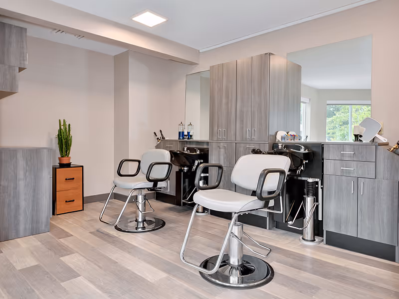 Interior view of a modern hair salon area within a senior living community, featuring two salon chairs in front of black wash basins, large mirrors, gray wooden cabinetry, and a small potted cactus on a wooden drawer unit.