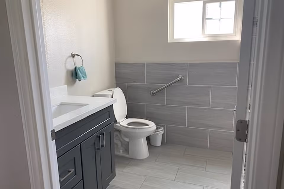 A clean, modern bathroom with gray tiled floor and walls halfway up. There is a white toilet with the lid open, a small trash can beside it, and a dark gray vanity with a white countertop and sink. A towel ring with a blue hand towel is mounted on the wall. A window above the toilet lets in natural light.