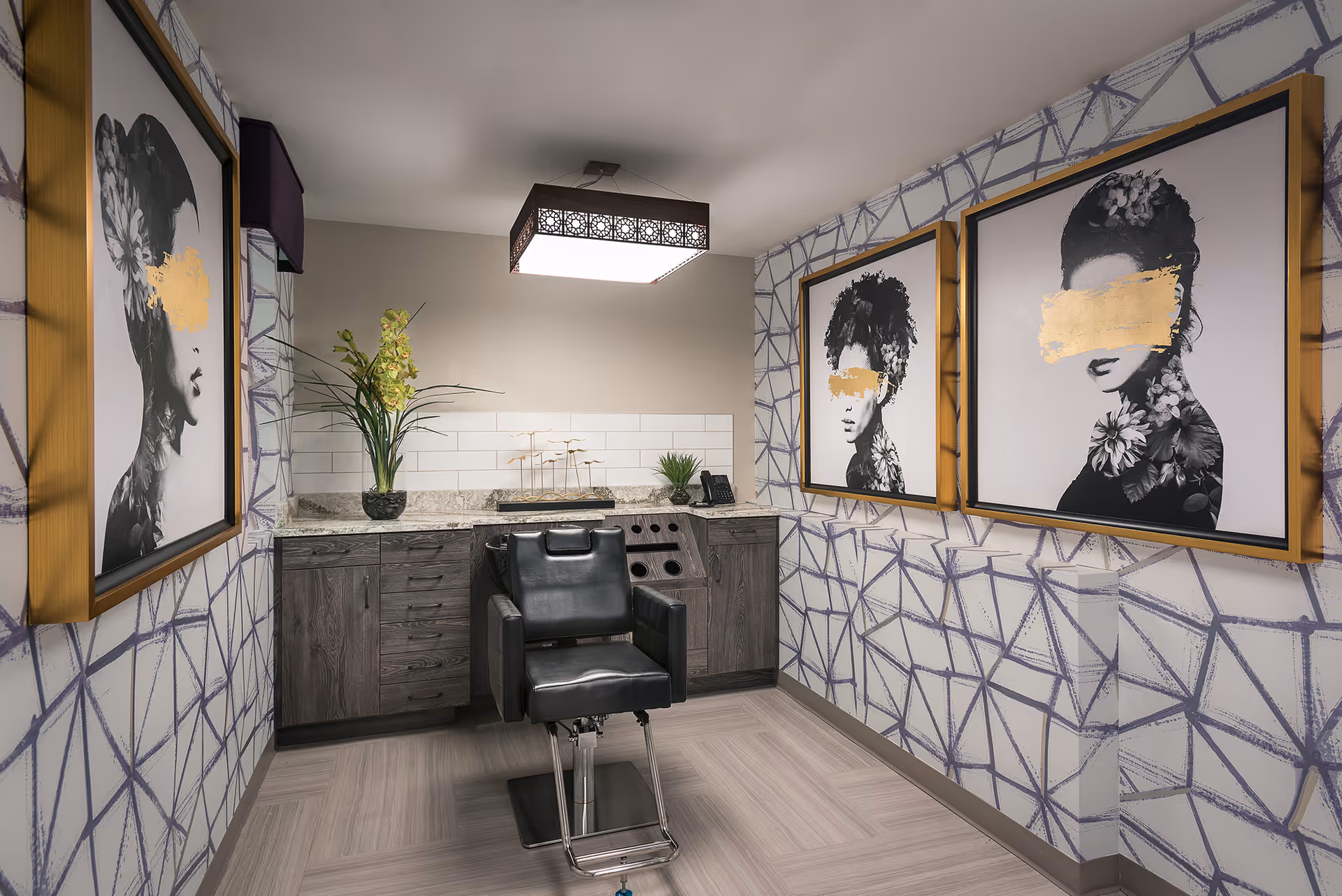 A modern salon room with a black salon chair in the center, dark wooden cabinets with a granite countertop, decorative plants, and three framed black and white portraits of women with gold paint obscuring their eyes on the walls. The walls have a geometric patterned wallpaper, and a rectangular ceiling light fixture illuminates the room.