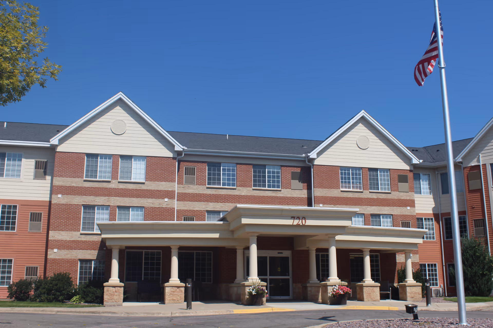 Three-story brick and siding senior living facility front entrance with a covered porte-cochère supported by columns and an American flag nearby.