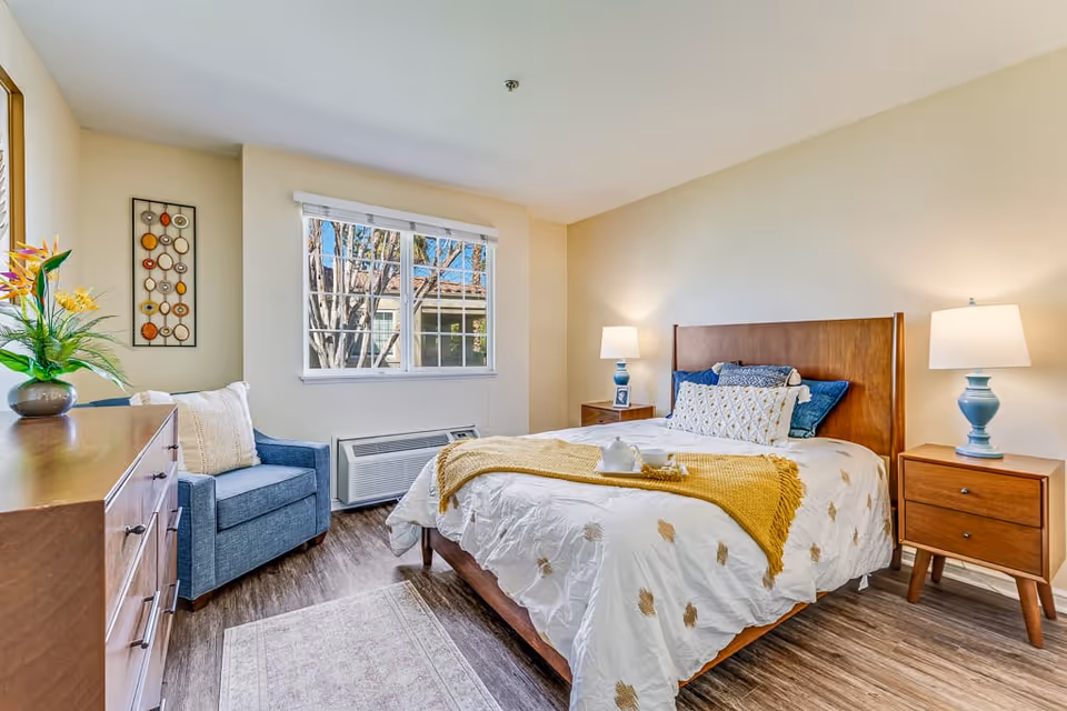 A bright and cozy bedroom with a wooden bed frame, white bedding with gold accents, and a yellow throw blanket. There are two wooden nightstands with blue lamps on either side of the bed. A blue armchair with a white pillow sits next to a window with white blinds. A wooden dresser with a vase of flowers is on the left side of the room. The floor is wood with a small area rug near the bed.
