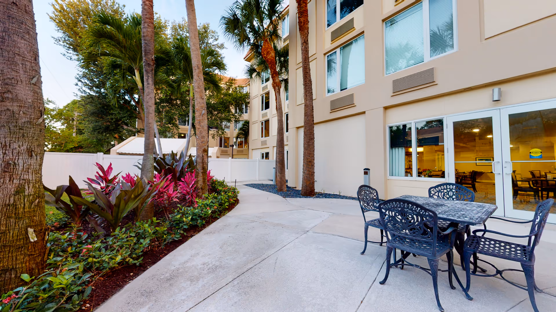 Outdoor patio area at The Atrium at Boca Raton with a metal table and four chairs on a concrete surface. The patio is adjacent to a building with large windows and glass doors. There are tall palm trees and landscaped plants with green and purple foliage along a white fence.