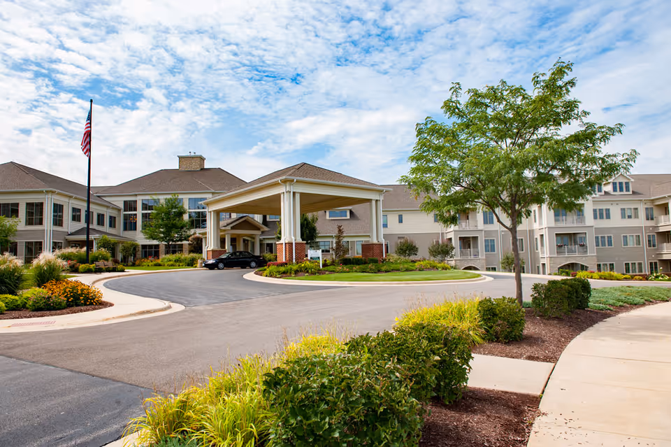 Front entrance of a senior living facility with a covered drop-off, circular driveway, landscaped grounds and an American flagpole.