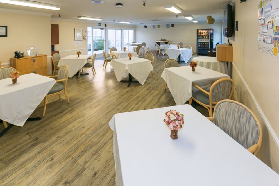 A dining room with several tables covered in white tablecloths, each adorned with a small floral centerpiece. The room has wooden flooring, beige walls, and multiple chairs around the tables. There is a vending machine and a television mounted on the wall in the background, with large windows and glass doors letting in natural light.