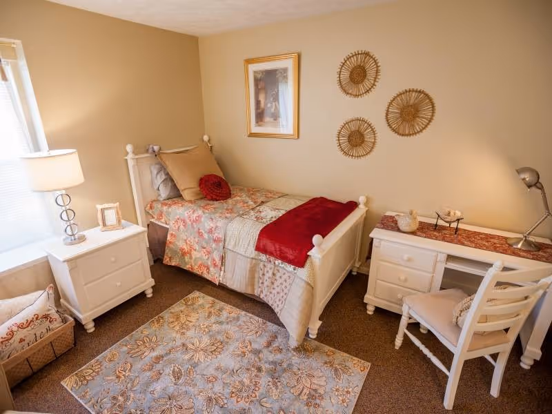 A cozy bedroom with a single white bed adorned with floral and patchwork bedding, a red throw blanket, and decorative pillows. Next to the bed is a white nightstand with a lamp and a small picture frame. On the opposite side, there is a white desk with a chair, a desk lamp, and decorative items. The walls are beige with three circular woven wall hangings and a framed picture. A patterned area rug covers part of the carpeted floor, and a basket with pillows is placed near the window.