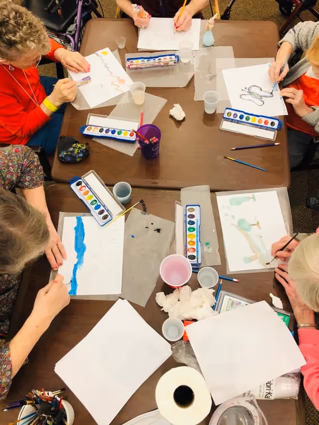 Overhead view of a group of elderly individuals seated around a table engaged in watercolor painting activities. Each person has a set of watercolor paints, brushes, and paper in front of them. The table also has cups of water, paper towels, and other painting supplies scattered around.