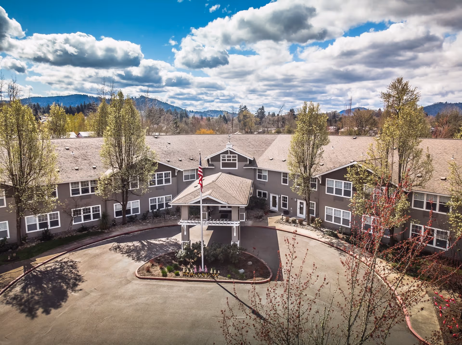 Exterior view of a senior living facility building with a circular driveway and a covered entrance. The building is two stories tall with multiple windows and surrounded by trees. The sky is partly cloudy with mountains visible in the background.