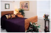 A bedroom with a wooden bed frame and a dark purple bedspread. Two decorative pillows with dog images are on the bed. There is a wooden nightstand with a lamp and a plant next to the bed. A framed picture hangs on the wall above the nightstand. A window with white blinds is on the right side, and a small round table with a flower arrangement is positioned in front of the window. Additional plants are placed near the foot of the bed.
