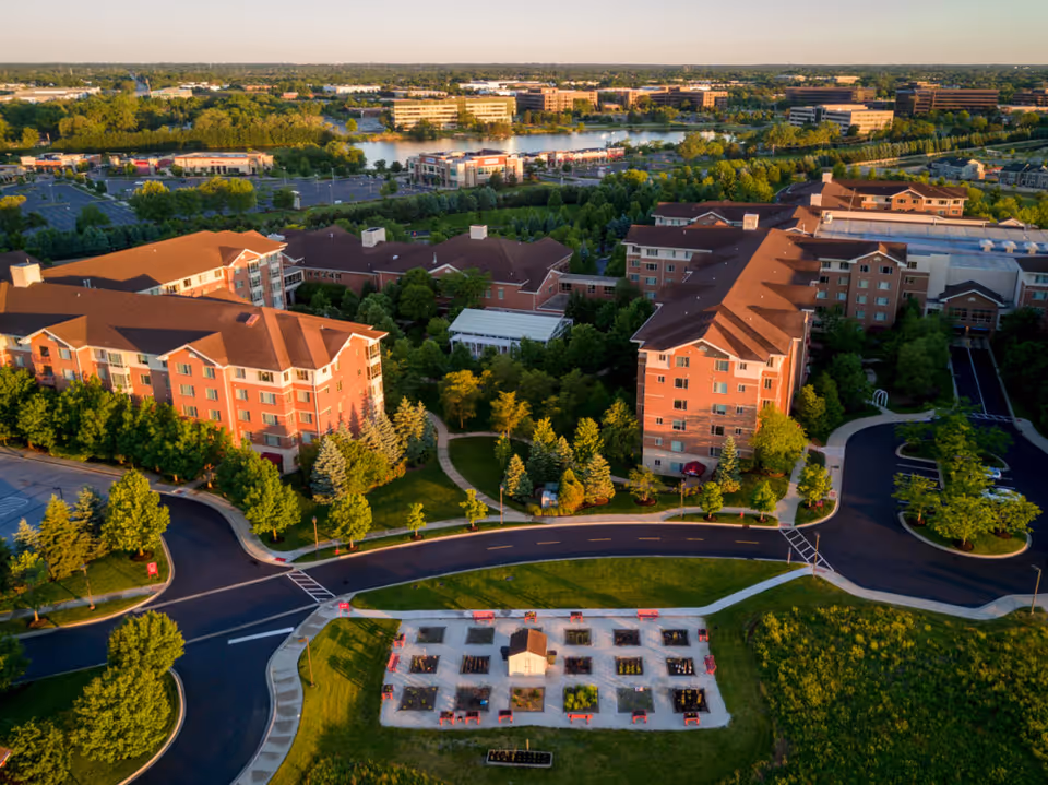 Aerial view of a senior living campus with multiple brick residential buildings, landscaped grounds, roads and a community garden.