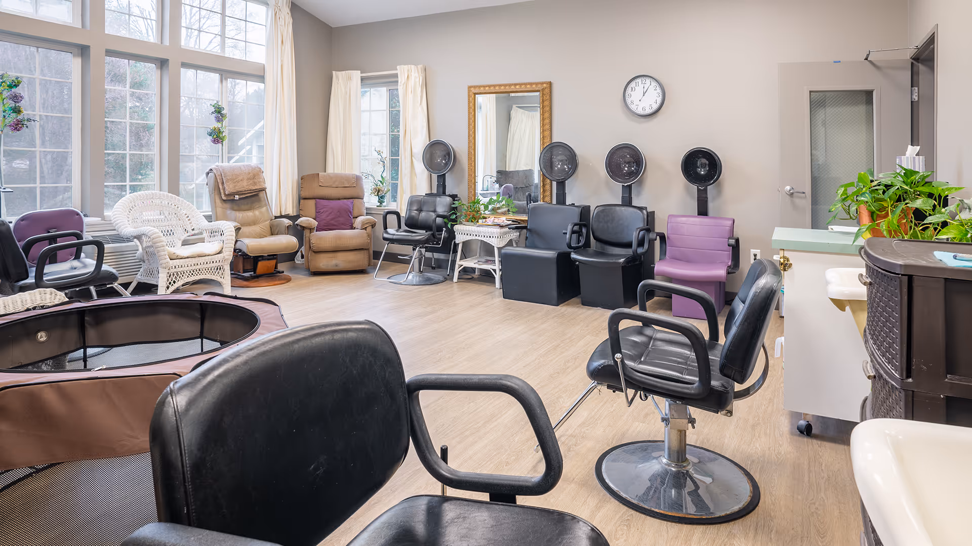 Interior view of a salon area in a senior living facility with multiple salon chairs, hair drying stations, comfortable armchairs, large windows with white curtains, a wall clock, and some green plants on a counter.