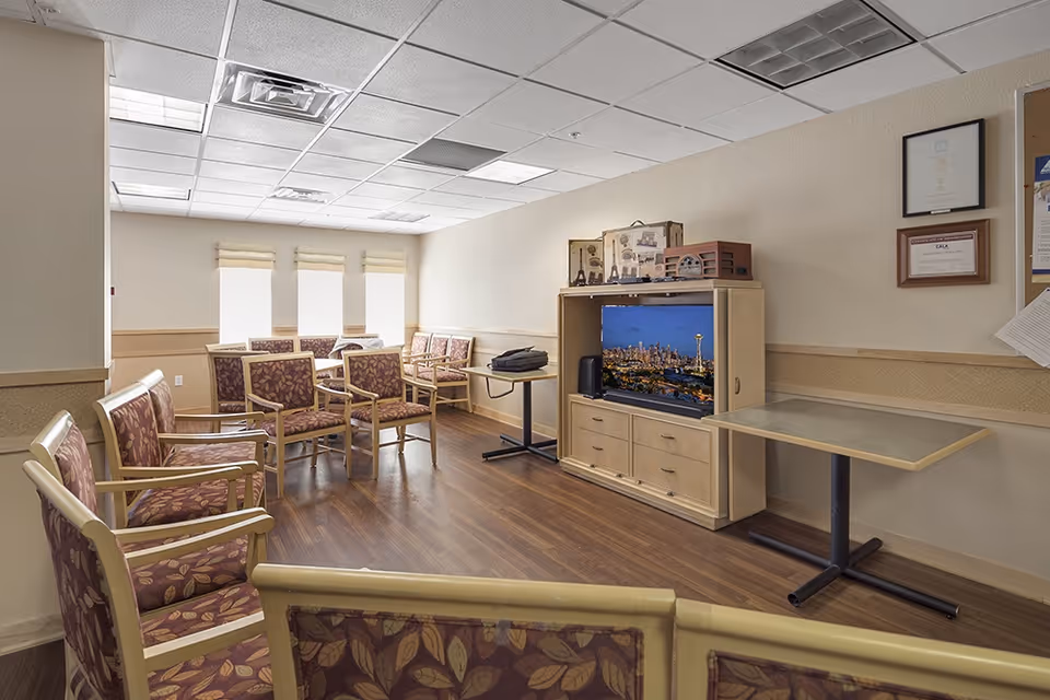 A common area in a senior living facility with multiple chairs arranged in rows and around tables. There is a wooden cabinet with a television displaying a cityscape, and some decorative items on top. The room has wood flooring, beige walls, and three windows with blinds partially drawn.
