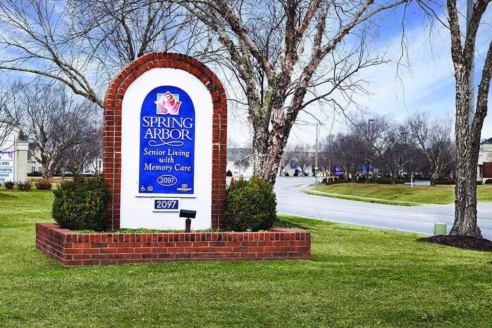 Brick-framed entrance sign reading "Spring Arbor Senior Living with Memory Care" on a grassy lawn with trees and a road in the background.