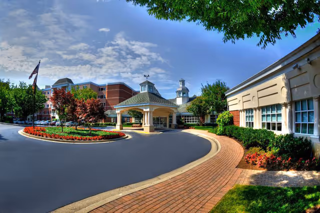 Exterior view of a senior living facility named Blakehurst, showing a circular driveway with a landscaped island of red flowers and trees, a covered entrance with a cupola, and multi-story brick and beige buildings under a partly cloudy sky.
