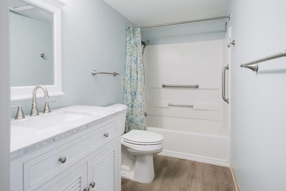A clean, modern bathroom with light blue walls, a white vanity with marble countertop and silver faucet, a toilet, and a white bathtub with a floral shower curtain and grab bars.