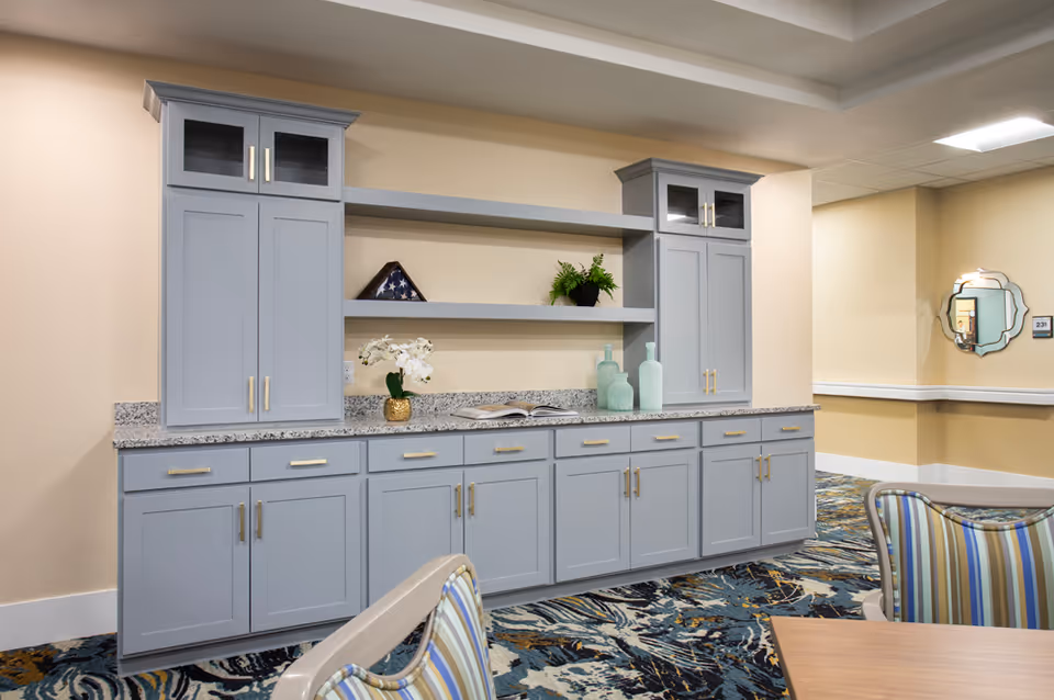 Interior view of a senior living facility hallway featuring a built-in cabinet with gray doors and gold handles, granite countertop, decorative items including a folded American flag, potted plants, and glass bottles. The floor has a patterned carpet and there are striped chairs and a wooden table partially visible. A decorative mirror and room number 231 are visible on the wall in the background.