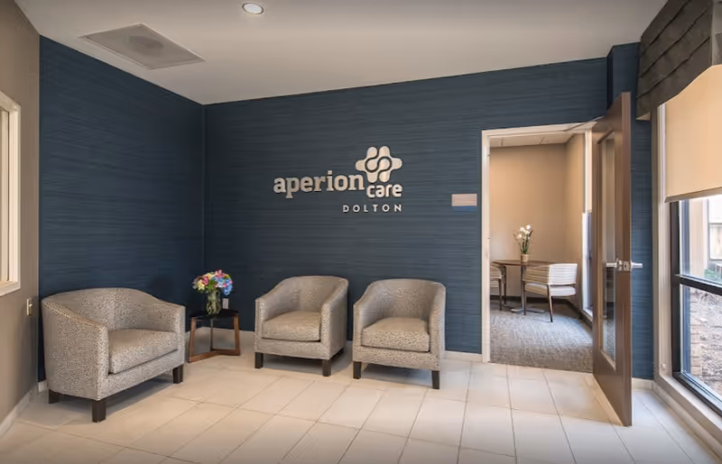 A waiting area with three gray upholstered armchairs arranged against a dark blue textured wall featuring the Aperion Care Dolton logo. A small wooden side table with a vase of colorful flowers is placed between two of the chairs. To the right, an open door leads to another room with a table, chairs, and a vase with white flowers. Large windows on the right side let in natural light.