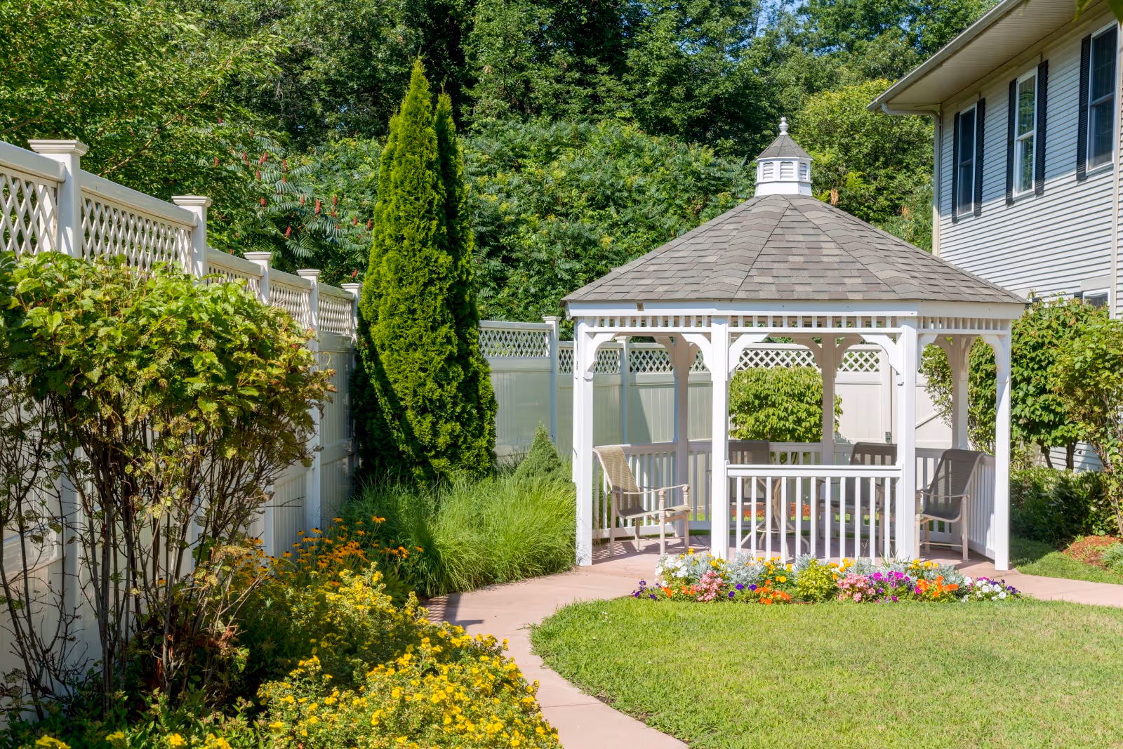 A white gazebo with a shingled roof in a garden area surrounded by green bushes, colorful flowers, and a white fence. There are chairs inside the gazebo and a paved walkway leading to it. A building with blue siding and windows is visible on the right side.