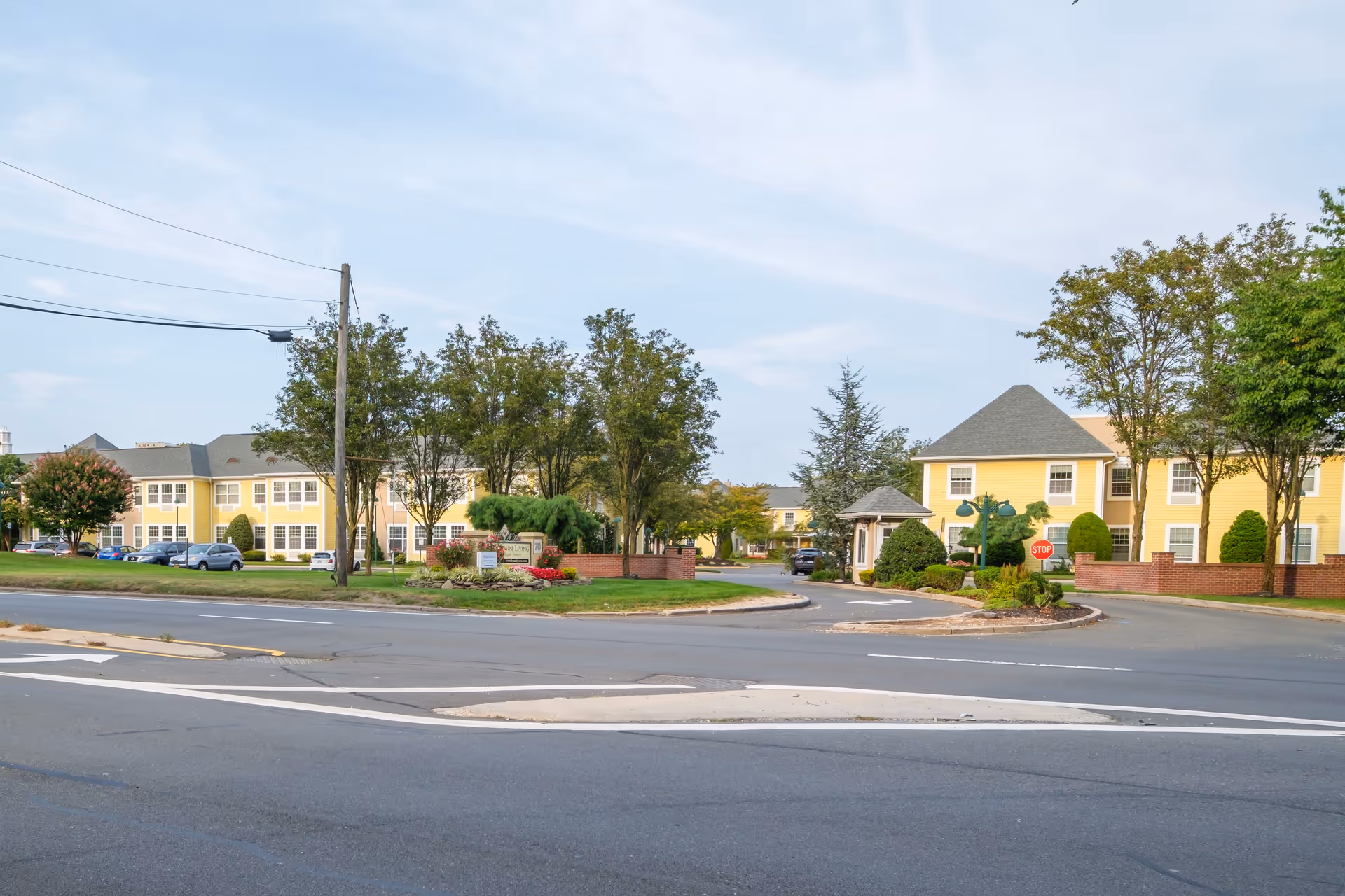 Exterior view of Brandywine Huntington Terrace by Monarch, showing yellow two-story buildings with gray roofs, surrounded by trees and landscaping, with a driveway entrance and a stop sign visible.