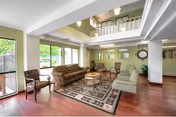 A spacious living room area in Woodbridge Manor featuring a patterned area rug, two sofas, two armchairs, a round wooden coffee table, and large windows letting in natural light. The room has hardwood floors, light-colored walls, and a high ceiling with a balcony railing visible above.