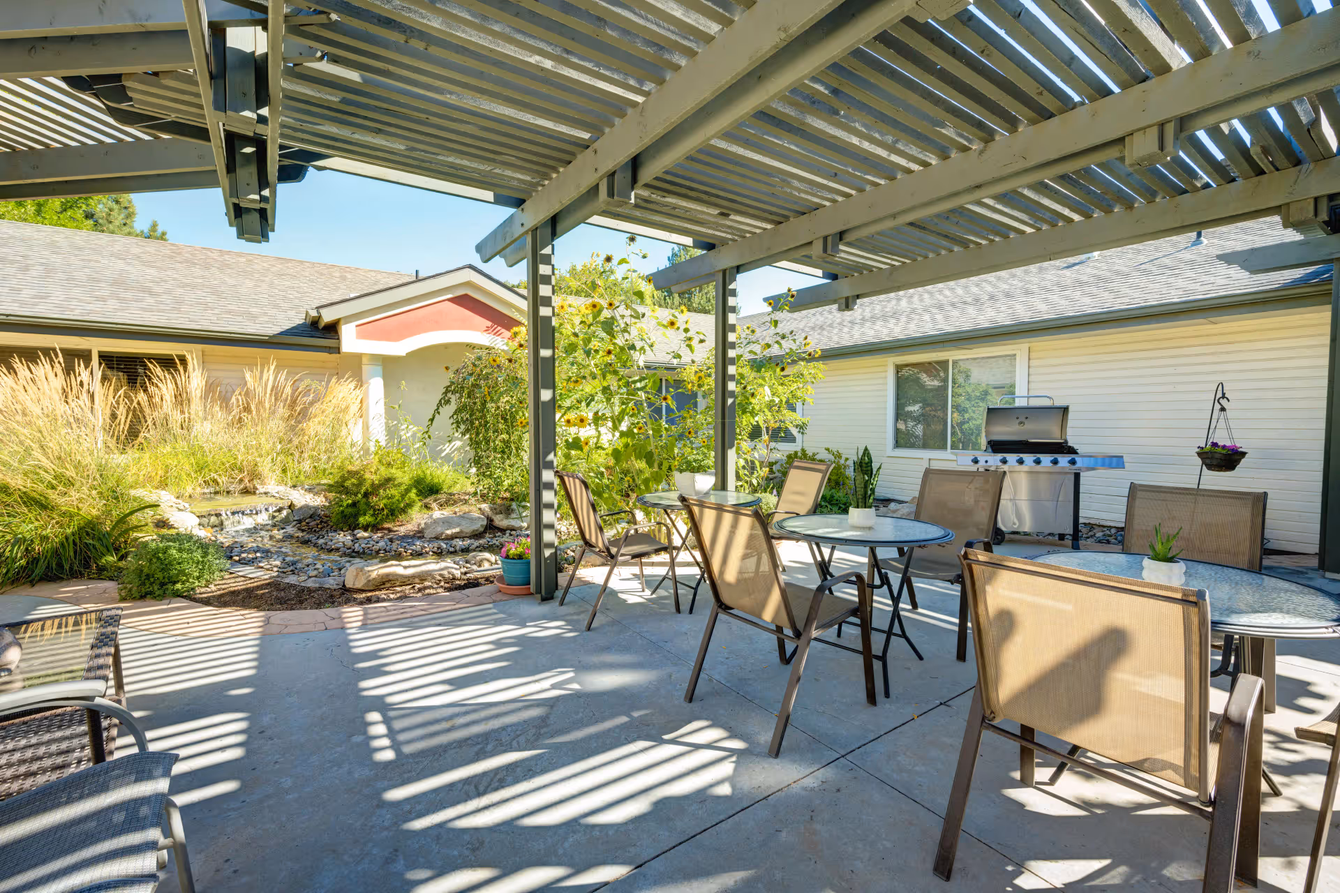 Shaded outdoor patio with tables and chairs, a grill, and landscaping under a pergola in a courtyard.