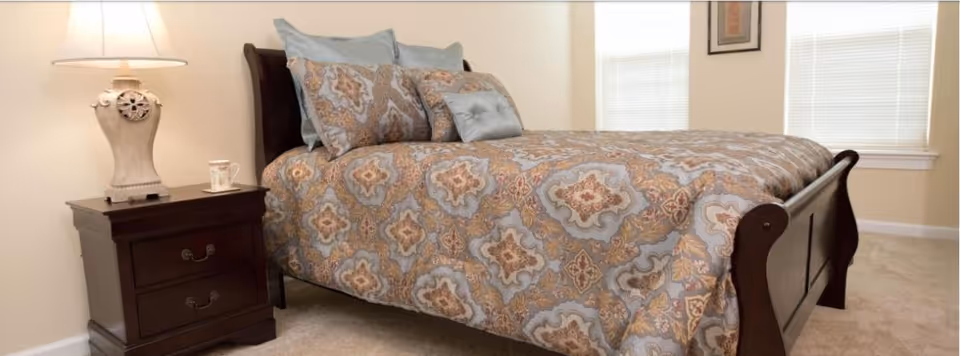Bright bedroom with a wooden sleigh-style bed dressed in a patterned comforter, a nightstand with a lamp and cup, and windows in the background.