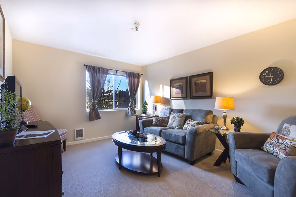A cozy living room with a large window covered by brown curtains, a gray sofa and armchair with decorative pillows, two table lamps on side tables, a round wooden coffee table with decorative items, a wall clock, framed artwork, and a TV on a wooden cabinet.