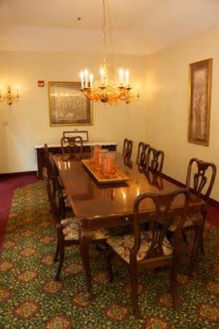 A formal dining room with a long wooden dining table surrounded by eight matching chairs with cushioned seats. A decorative chandelier with lit candles hangs above the table. The room has patterned carpet flooring and light-colored walls adorned with framed artwork and wall sconces.