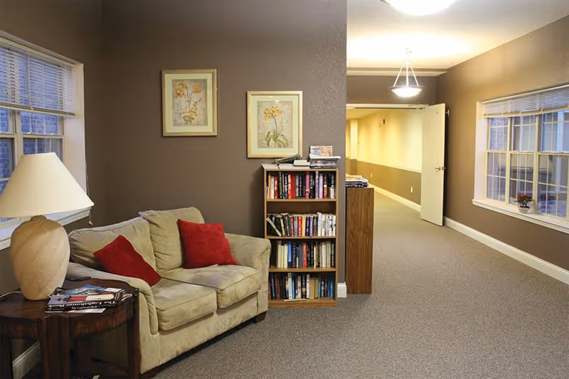 A cozy seating area in a hallway with a beige loveseat adorned with two red pillows, a wooden side table with a lamp and magazines, a wooden bookshelf filled with books, two framed floral paintings on the wall, and windows on both sides letting in natural light.
