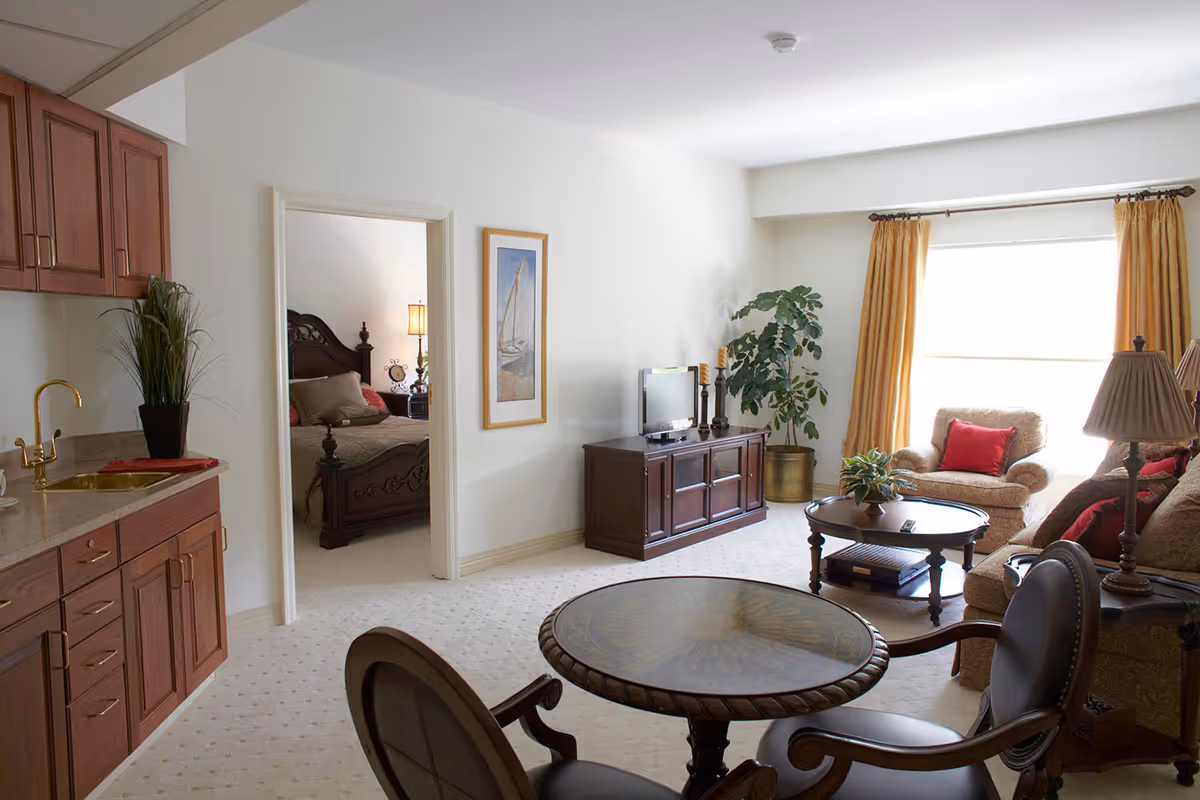 A bright living room with beige carpet, a round wooden table with two chairs in the foreground, a wooden TV stand with a flat screen TV, a potted plant, and a seating area with a sofa and armchair with red cushions near a large window with yellow curtains. To the left, there is a kitchenette with wooden cabinets and a gold faucet. An open doorway reveals a bedroom with a dark wooden bed frame and a bedside lamp.