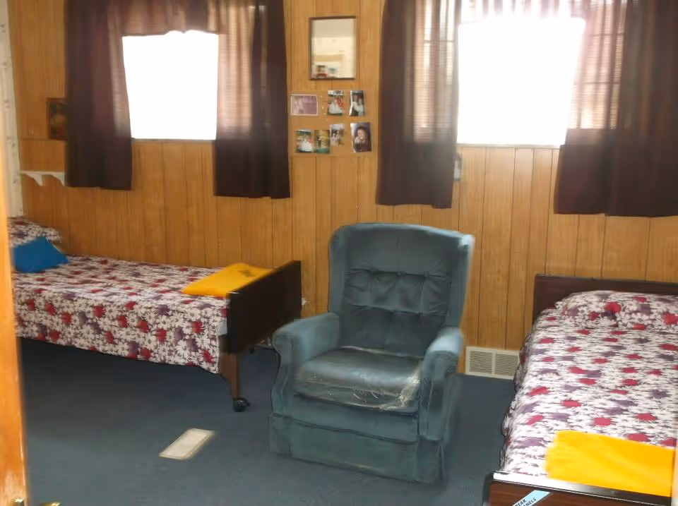 Small room with two single beds and a green upholstered recliner between them, wood-paneled walls, curtained windows, and photos on the wall.