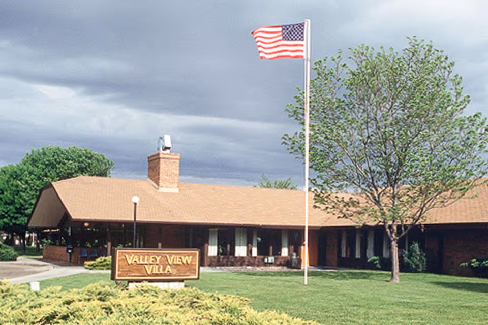 Exterior view of Valley View Villa, a single-story building with a tan roof and brick walls. There is a green lawn in front with a tree and an American flag on a flagpole. A wooden sign with the facility name is visible near the entrance.