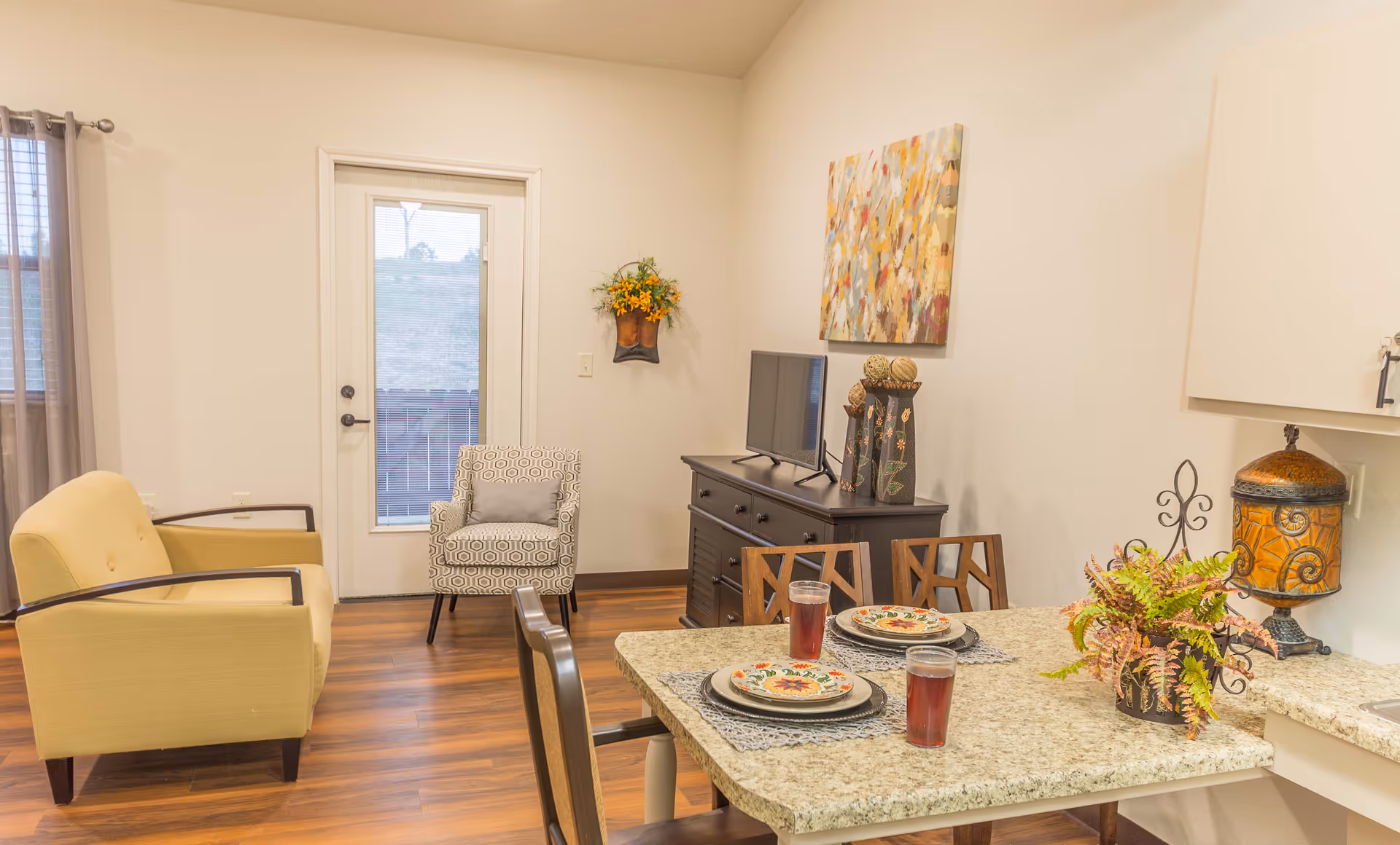 A cozy living and dining area in an assisted living facility featuring a yellow armchair, a patterned armchair, a small TV on a dark wooden dresser, a dining table set with plates and glasses of red beverage, and decorative plants and artwork on the walls.
