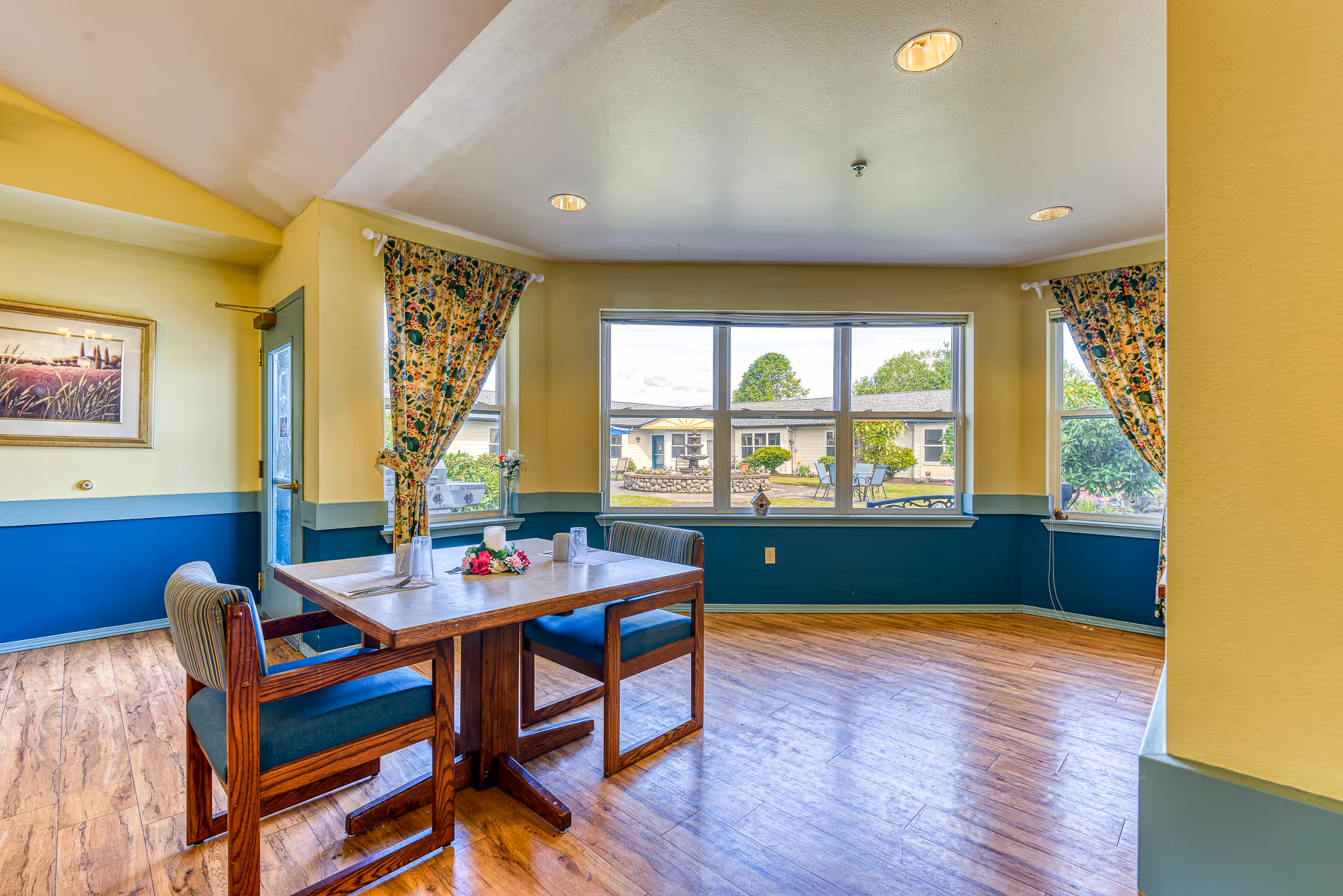 A bright dining area with wooden flooring, a wooden table with two chairs featuring blue cushions, and large windows showing an outdoor garden with a fountain and seating. The walls are painted yellow with a blue lower half, and floral curtains frame the windows.