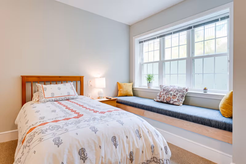 A bright bedroom featuring a single wooden bed with patterned white and red bedding, a wooden nightstand with a lamp, and a large window seat with blue cushion and decorative pillows beneath a multi-pane window.